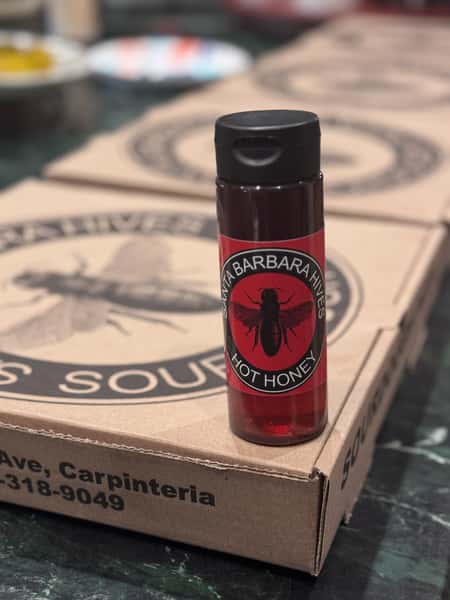 Bottle of Santa Barbara Hives Hot Honey with a red label and bee logo, placed on branded cardboard pizza boxes.