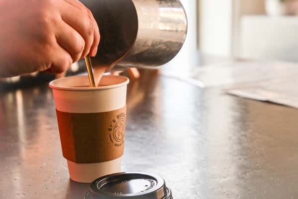 Hand pouring warm drink from metal pitcher into AJ's Riverside paper cup with brown sleeve.