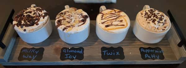 Four candy-themed hot chocolates (Milky Way, Almond Joy, Twix, Peppermint Patty) with whipped cream and toppings on a gray tray from AJ's Riverside.