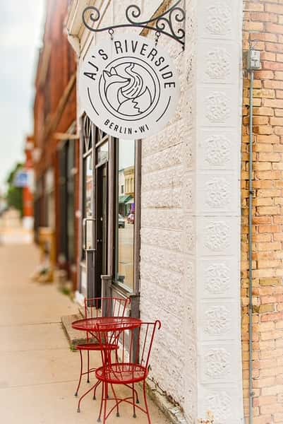 AJ's Riverside white sign with fox logo on a building exterior wall beside red outdoor bistro chairs and table.
