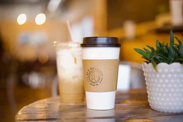 AJ's Riverside hot coffee cup with logo sleeve and iced coffee on a wooden table with a plant.