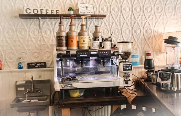 AJ's Riverside coffee bar featuring a silver espresso machine, grinders, various syrup bottles, and white textured walls.