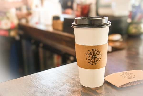 White paper coffee cup with AJ's Riverside logo sleeve on a cafe counter.