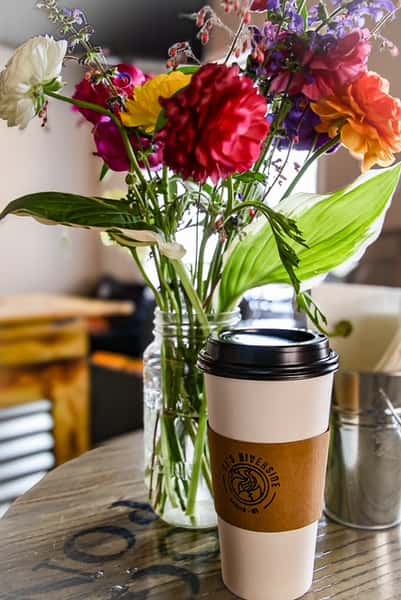 White AJ's Riverside coffee cup with colorful bouquet of flowers on a wooden table.