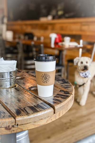 AJ's Riverside logo coffee cup on a rustic wooden table in a cafe with a dog in the background.