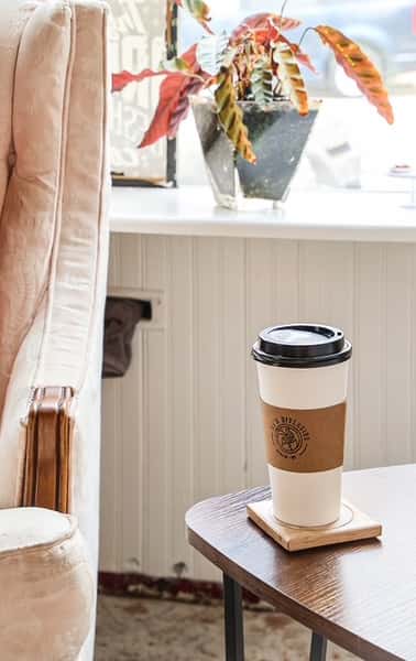 White coffee cup with AJ's Riverside logo on brown sleeve, on wooden table next to plush armchair.