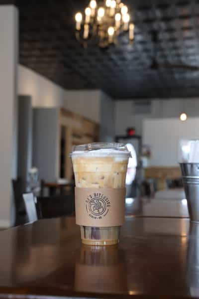 Creamy iced coffee in a cup with AJ's Riverside logo sleeve on a table.