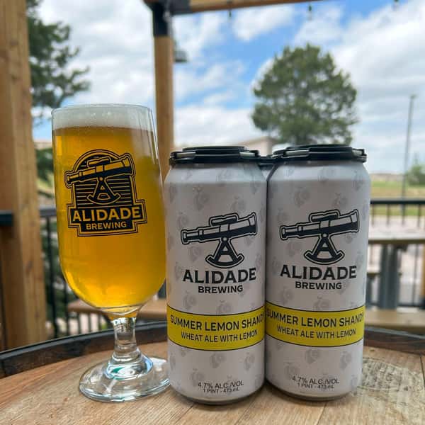 Glass of Alidade Brewing Summer Lemon Shandy served alongside cans of the seasonal wheat ale with lemon on the brewery's outdoor patio.