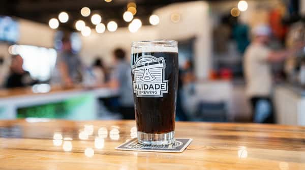 Dark beer in a glass with the "Alidade Brewing" logo, on a wooden bar in a brewery.