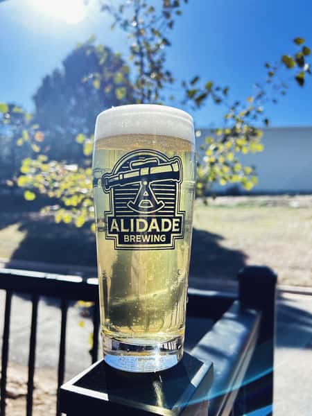 Crisp golden craft beer in an Alidade Brewing pint glass enjoyed outdoors on the patio, showcasing a refreshing pour with a bright foamy head.