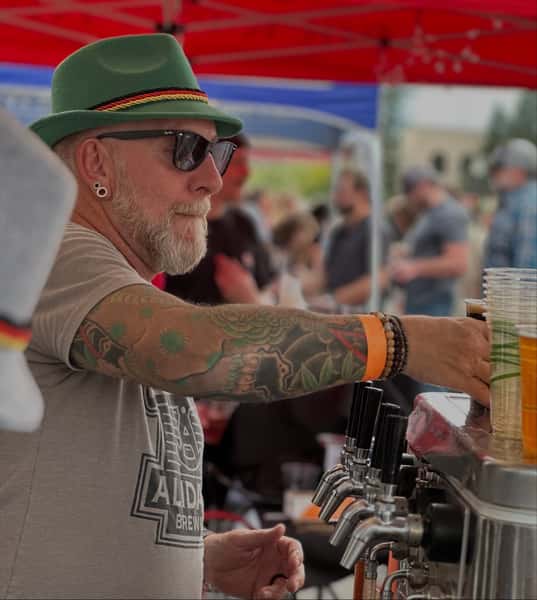 Beer festival guest pouring and serving craft beer at an Alidade Brewing tap station during a lively outdoor community event.