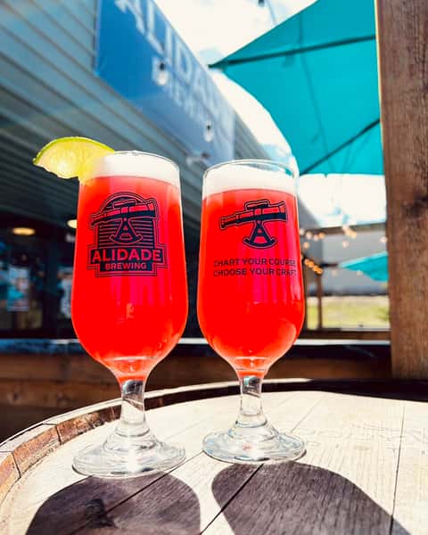 Two vibrant fruit-forward craft beers in Alidade Brewing glasses served on the outdoor patio, featuring a bright red pour, fresh lime garnish, and sunny brewery atmosphere.