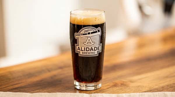 Dark craft beer in an Alidade Brewing pint glass featuring a rich caramel-colored head, served on a wooden bar top and showcasing the brewery's signature stout or porter style.