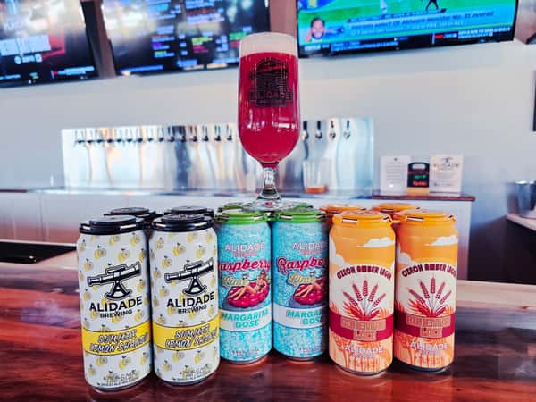 Alidade Brewing canned craft beer lineup featuring Summer Lemon Shandy, Raspberry Lime Margarita Gose, and Bohemian Lace Czech Amber Lager displayed in the brewery taproom with a vibrant fruit beer pour.