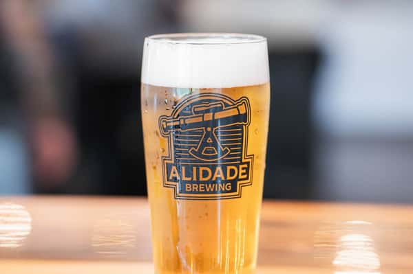 Crisp golden craft lager in an Alidade Brewing pint glass featuring a bright pour, refreshing foam head, and the brewery's signature logo.