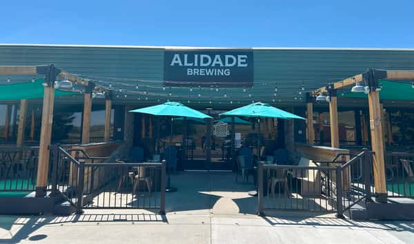 Front entrance of Alidade Brewing featuring a spacious outdoor patio, turquoise umbrellas, string lights, and welcoming brewery seating.