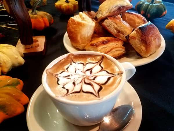 capuccino in a white cup and saucer with decorated foam. pastries on the table in the background.