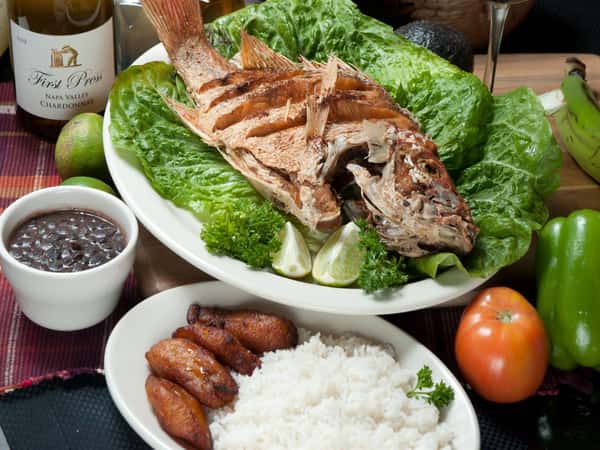 Pargo Entero Frito. Whole Fried Red Snapper on a bed of greens. White rice, fried plantains and black beans on the side. bottle of white wine, green pepper, tomato and avocado in the background.