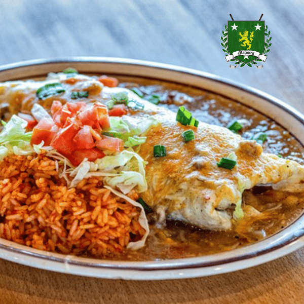 A hearty Mexican-style plate featuring a cheese-smothered entrée topped with diced tomatoes and green onions, served alongside seasoned Spanish rice. The dish is presented on a rustic brown platter, with melted cheese sauce covering most of the main item, creating a warm and comforting appearance. The colorful garnish adds freshness and contrast to the rich, savory meal.