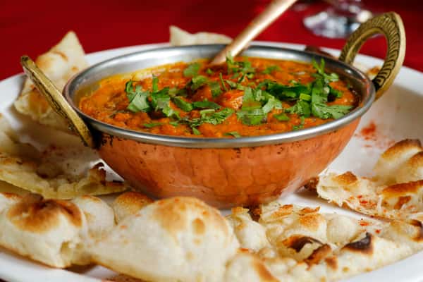 Rich Indian curry topped with fresh cilantro, served in a copper bowl with pieces of naan bread on a white plate.