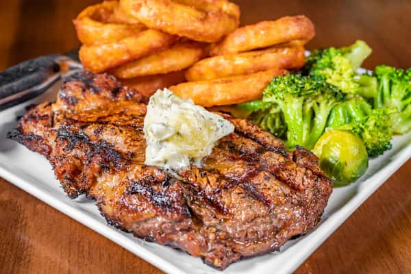 Grilled ribeye steak topped with herb butter, served with golden onion rings and fresh broccoli at Pourhouse Bar & Grill in Loveland.