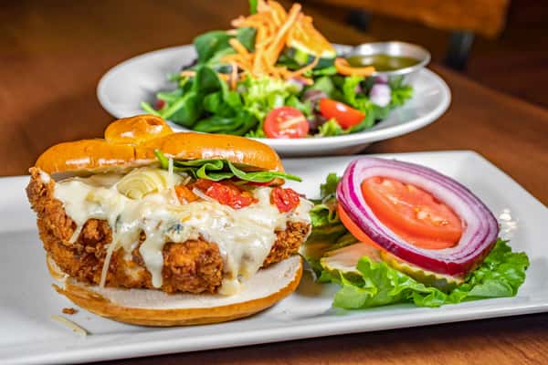 Crispy fried chicken sandwich topped with melted white cheese, roasted tomatoes, and fresh greens on a toasted brioche bun, served with lettuce, tomato, red onion, pickles, and a side salad at Pourhouse Bar & Grill in Loveland.