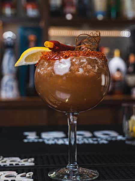 Smoky Mexican cocktail with chili rim, lemon wedge, and cinnamon garnish served at a modern tequila bar