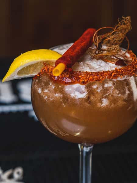 Spicy Mexican cocktail with chili rim, lemon garnish, and smoky cinnamon straw served over crushed ice
