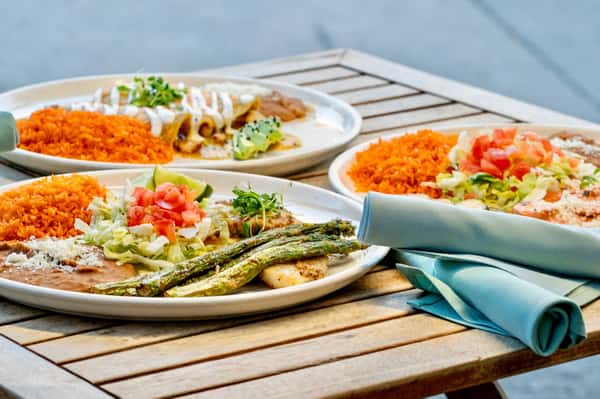 Colorful Mexican dinner plates with enchiladas, rice, refried beans, grilled vegetables, and fresh salad served on a rustic outdoor patio table