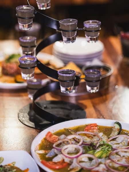 Tequila shot tower with salt-rimmed glasses beside aguachile seafood dish at a vibrant Mexican restaurant