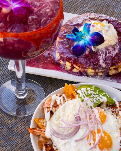 Purple brunch cocktail served with berry pancakes, chilaquiles topped with eggs and queso fresco, and colorful Mexican brunch dishes