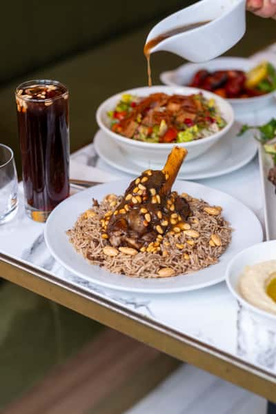 Rich lamb shank served over seasoned rice with toasted nuts and savory sauce alongside fresh Mediterranean salad and mezze