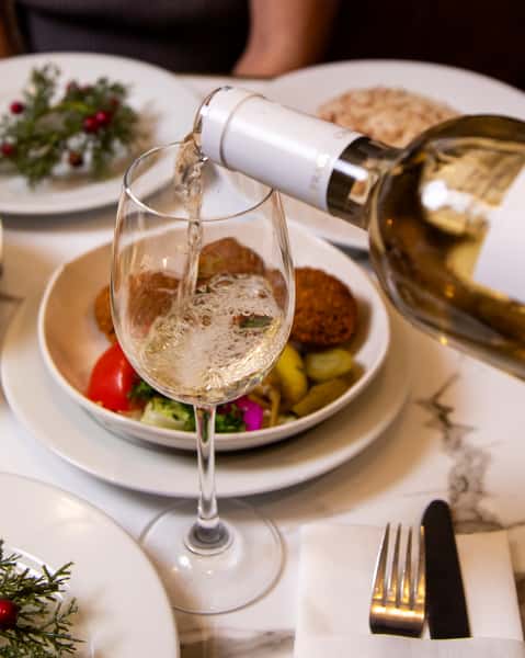White wine being poured into a glass beside fresh falafel and Mediterranean mezze on a marble dining table