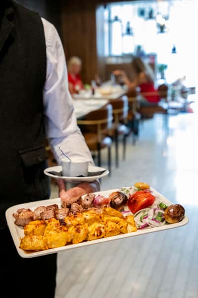 Server carrying a grilled mixed kebab platter with roasted vegetables and garlic sauce through a modern restaurant dining room