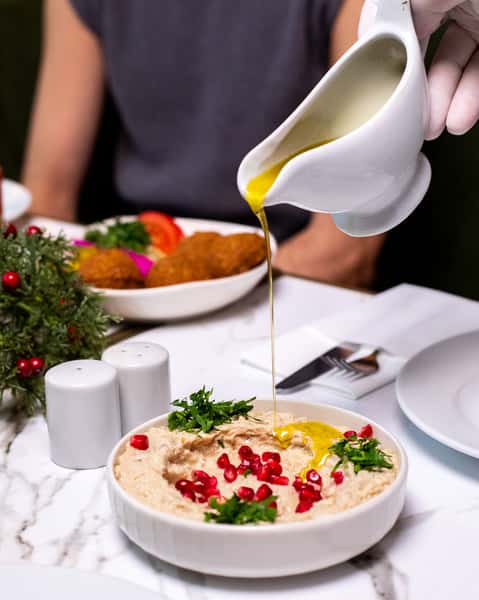 Olive oil being poured over creamy baba ghanoush topped with pomegranate seeds and fresh herbs at a Mediterranean restaurant