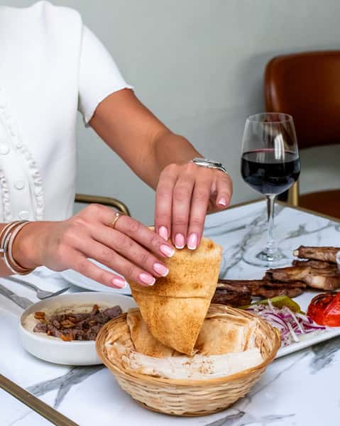 Guest dipping warm pita bread into creamy hummus served with grilled meats, red wine, and Mediterranean sides