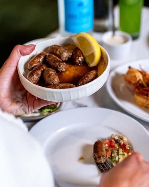 Hand holding a bowl of grilled Mediterranean sausages served in warm sauce with a lemon wedge at a modern Lebanese restaurant