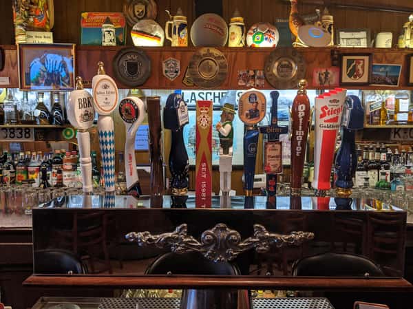 Row of colorful beer taps at a bar with German steins, plaques, and bottles displayed in the background.