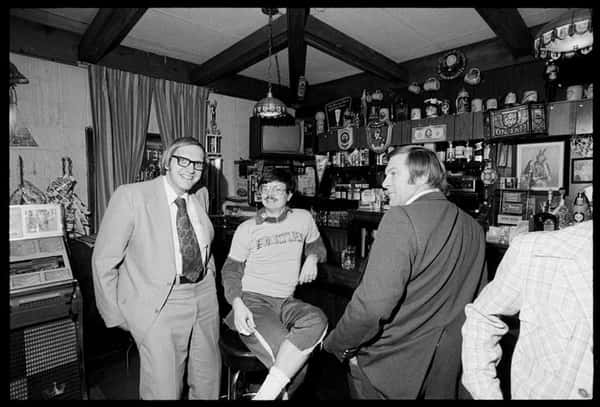 Three men socializing at a vintage bar, with one seated casually and two in suits, surrounded by steins and decor.