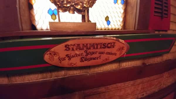 Wooden plaque with German text reading "Stammtisch für Fischer, Jäger und andere Lügner!" mounted below stained glass.