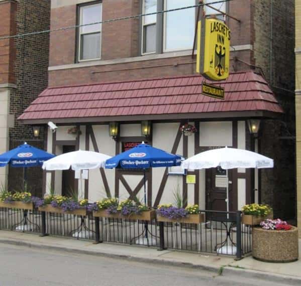 Laschet's Inn restaurant exterior with a yellow sign, red roof, and outdoor seating under umbrellas with flower planters.