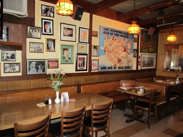 Cozy restaurant corner with wooden tables, framed photos, and a mural with German text on the wall.