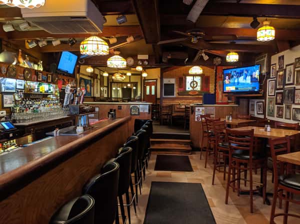 Cozy pub interior with a long bar, barstools, wooden tables, TVs, and warm stained-glass style hanging lights.
