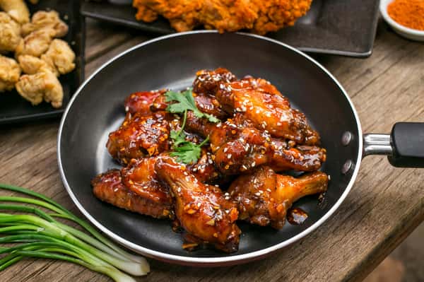 This image shows a pan filled with glazed chicken wings coated in a sticky, dark sauce topped with sesame seeds and garnished with a sprig of fresh cilantro. The wings appear to have a sweet and savory glaze, likely with garlic and chili flakes. In the background, there are pieces of fried chicken and green onions on the table.