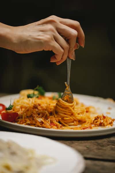 Hand twirling spaghetti with tomato sauce and grated cheese on a plate, garnished with cherry tomatoes and herbs.