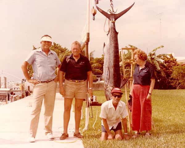 A group of four people posing near a large caught fish displayed on a pole.
