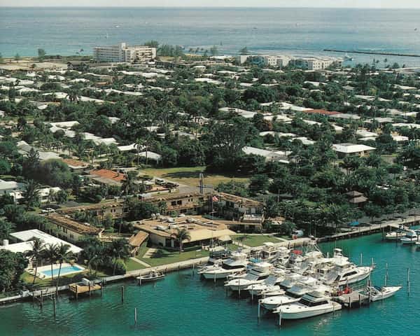 Aerial view of a lush coastal neighborhood with homes and a marina filled with boats, set against a blue ocean.