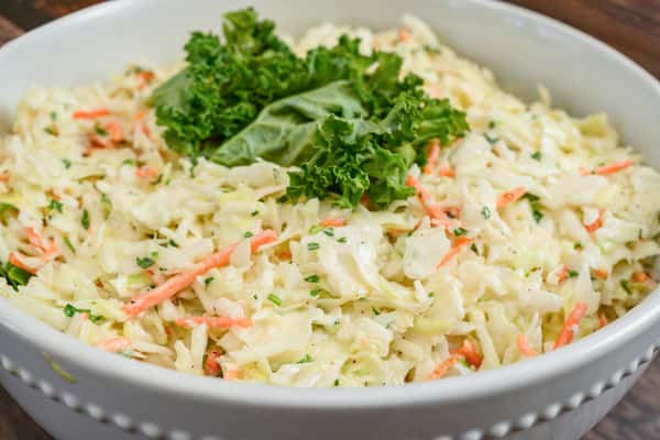 Creamy coleslaw made with shredded cabbage, carrots, and herbs, garnished with leafy kale on top, served in a white bowl.