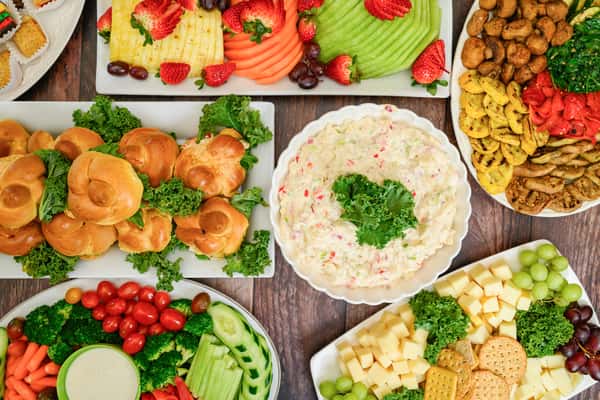 Colorful party spread with mini sandwiches, veggie platter, fruit slices, cheese cubes, and a creamy dip topped with kale.