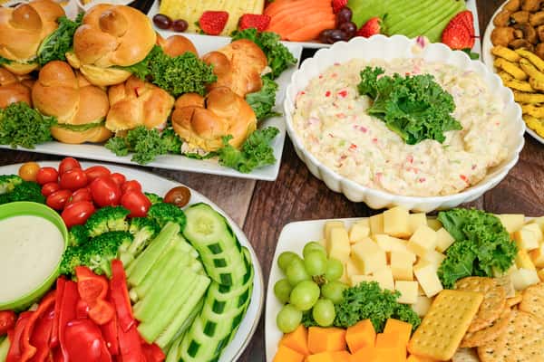 Assorted party platter with mini sandwiches, cheese cubes, potato salad, fresh vegetables, fruit, and dipping sauces.